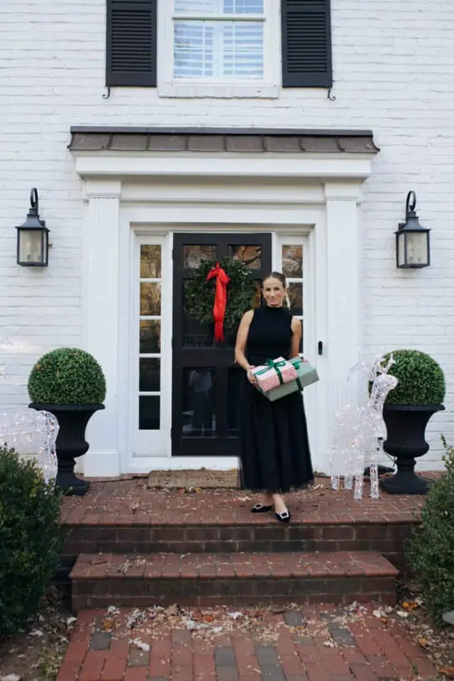 woman in black tulle skirt holding christmas gifts on porch in front of white house